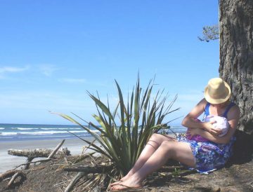 Mother sitting up against a tree by a West Coast beach.  She's holding her baby close to the breast to feed it.