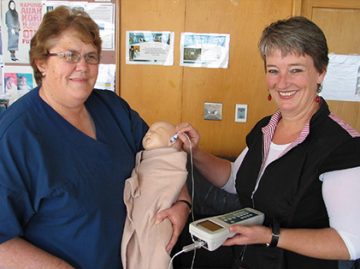 Hearing screeners Barbara Roberts and Mary Sullivan holding a plastic doll.  Hearing screening equipment is being held by the ear of the doll.